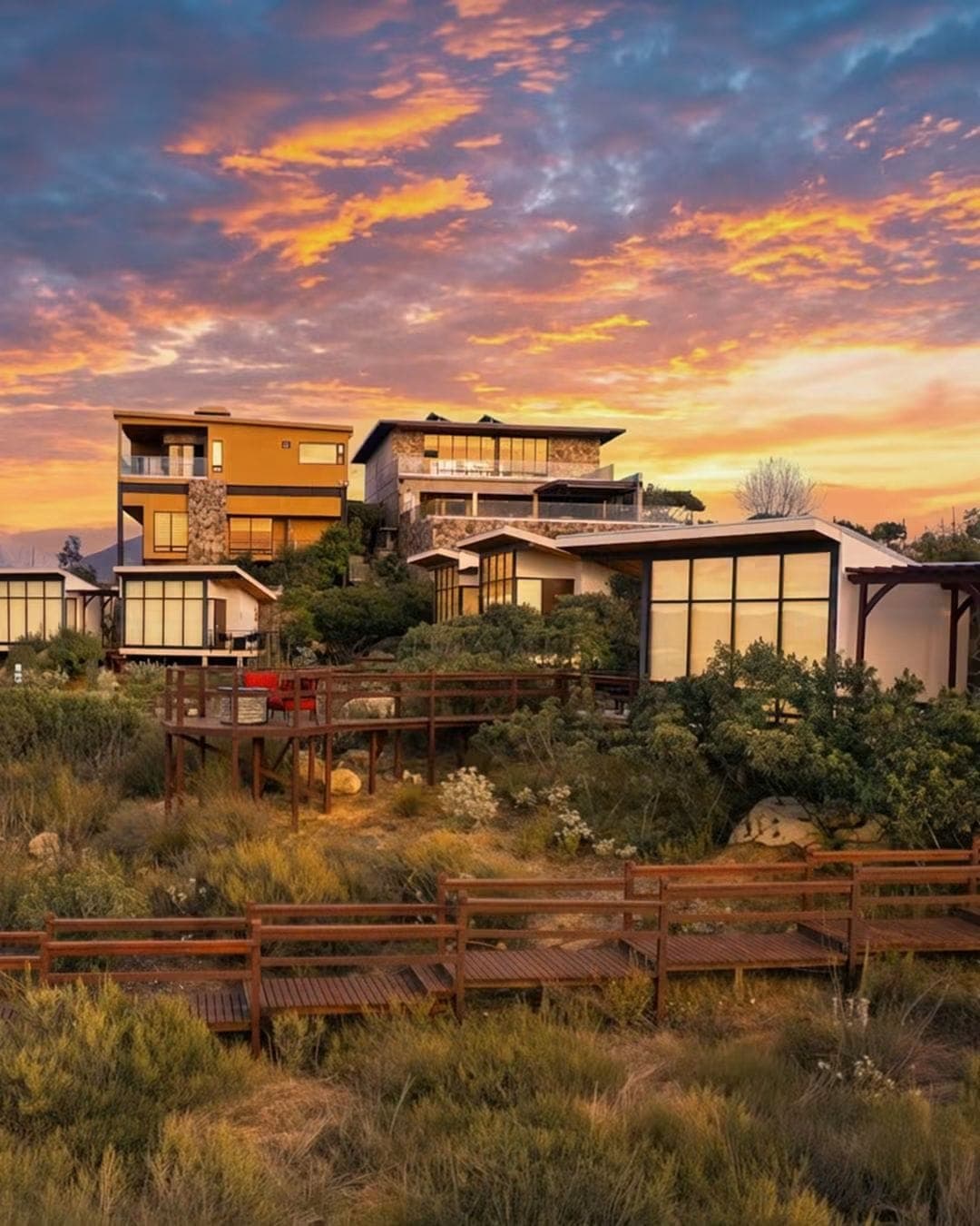 Cabañas de Contemplación bajo cielos rojizos del atardecer, con pasarelas de madera atravesando el paisaje natural